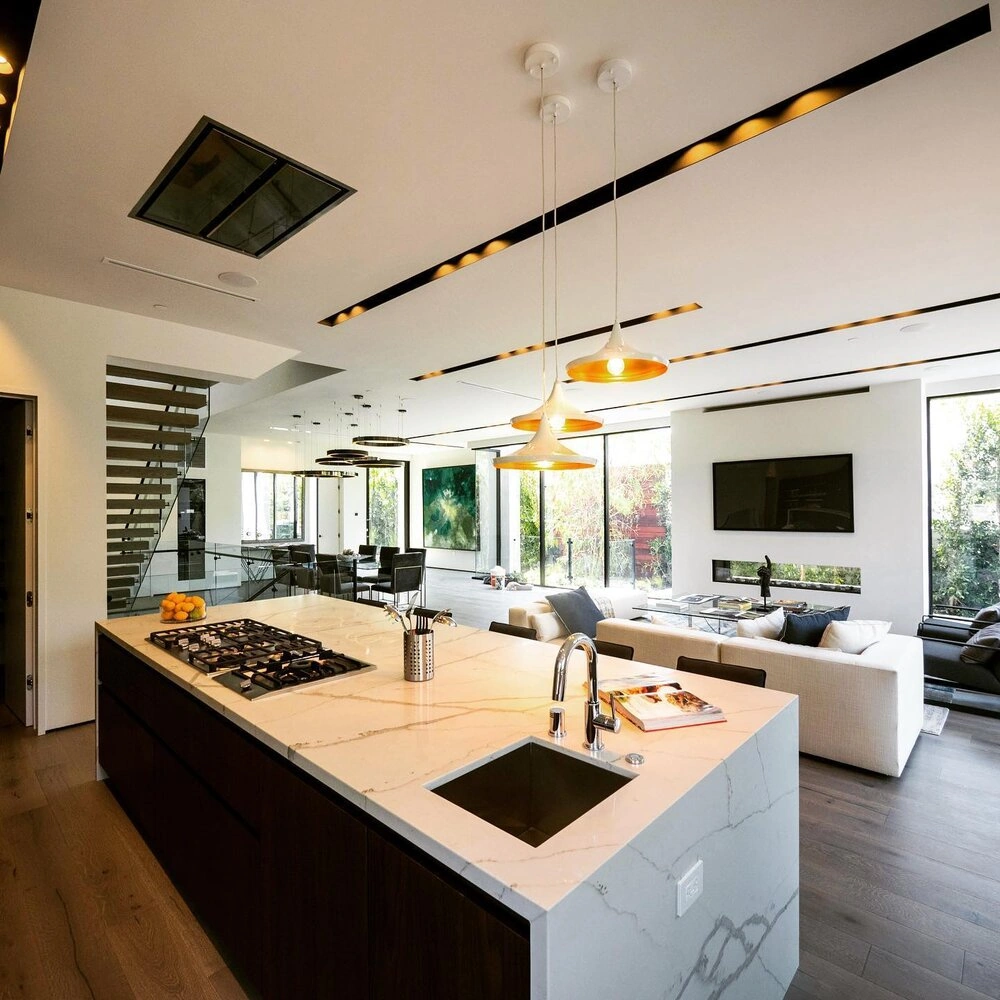 Open-concept kitchen with a waterfall marble island, built-in cooktop, and brass pendant lights flowing into a modern living room