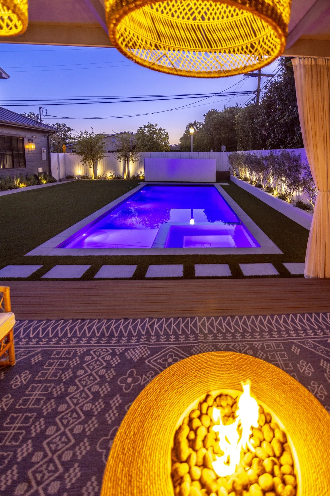 Custom pool with an attached spa and color LED lighting at night in Los Angeles, viewed from a pergola lounge with a fire pit