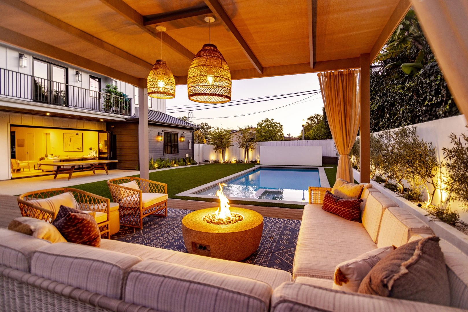 Pergola lounge in Los Angeles with woven pendant lights, an outdoor sectional, and a fire pit overlooking the backyard pool