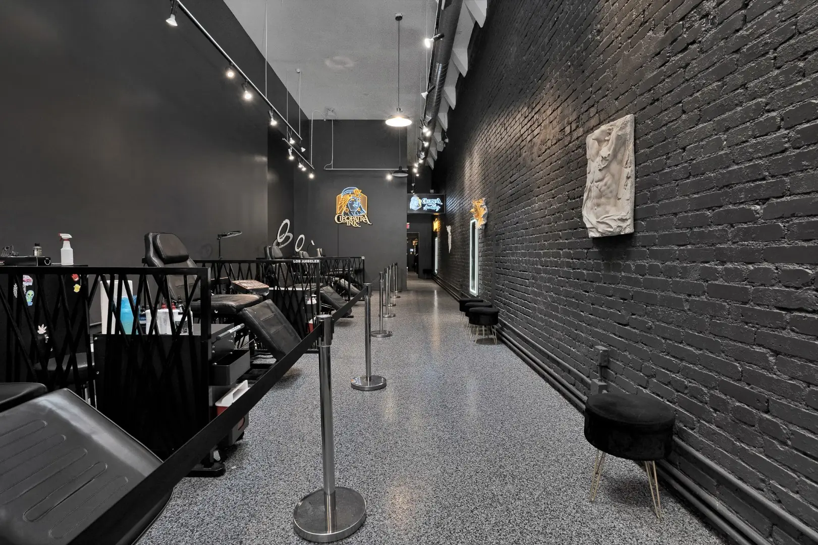 Cleopatra Tattoo stations along a painted black brick wall with track lighting in Santa Monica