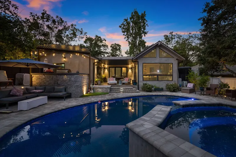 Freeform pool with a raised spa at dusk in Los Angeles, facing an outdoor kitchen under a louvered pergola, a lit paver patio, and a modern gabled home