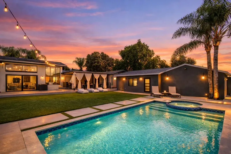 Rectangular pool with a round spa and blue tile waterline at dusk, beside concrete pavers set in turf, a curtained pergola, and a modern single-story home