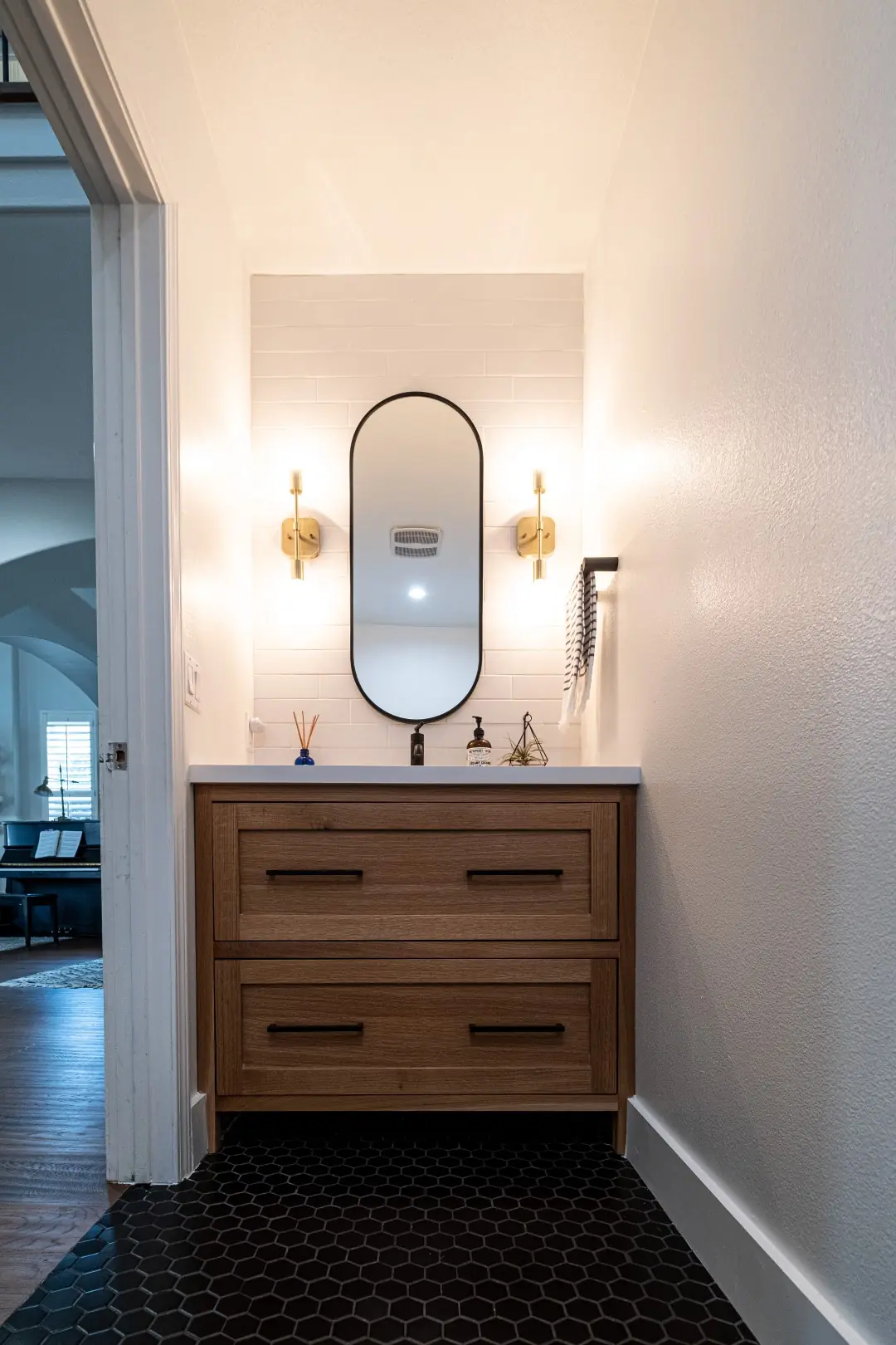 Powder room in a West LA renovation with a walnut vanity, oval black mirror, brass sconces, and black hexagon floor tile