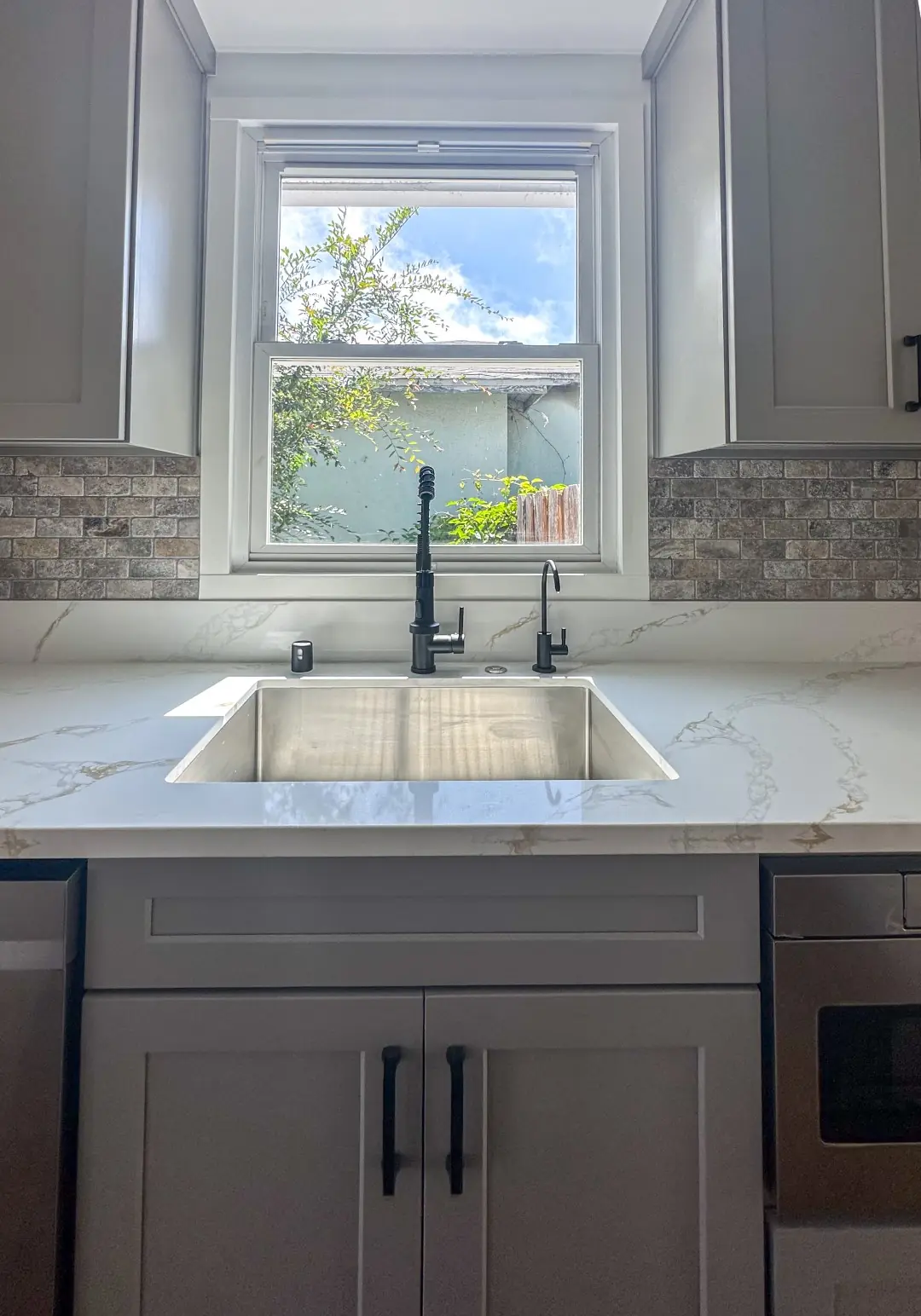Stainless sink and matte black faucet under a window with a stone mosaic backsplash in a Santa Monica kitchen