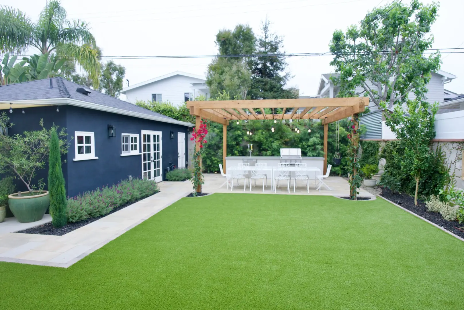 Agnew Project backyard in Los Angeles with an artificial turf lawn leading to a wood pergola, outdoor kitchen, and navy guest house