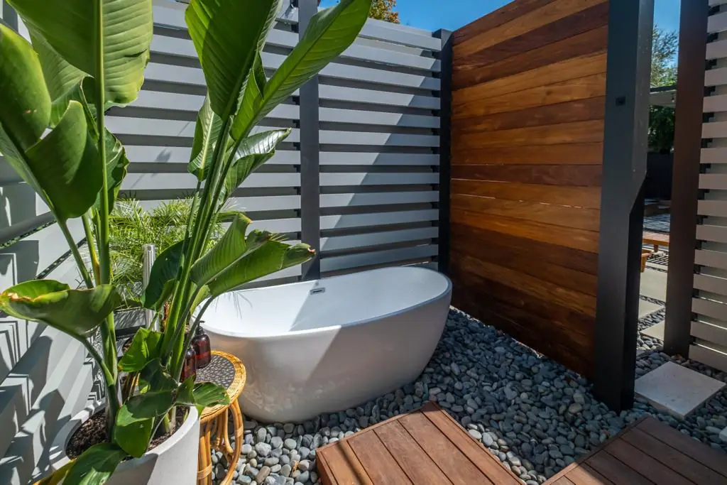 Outdoor freestanding soaking tub set in river pebbles behind a wood slat privacy screen with tropical plants in Los Angeles