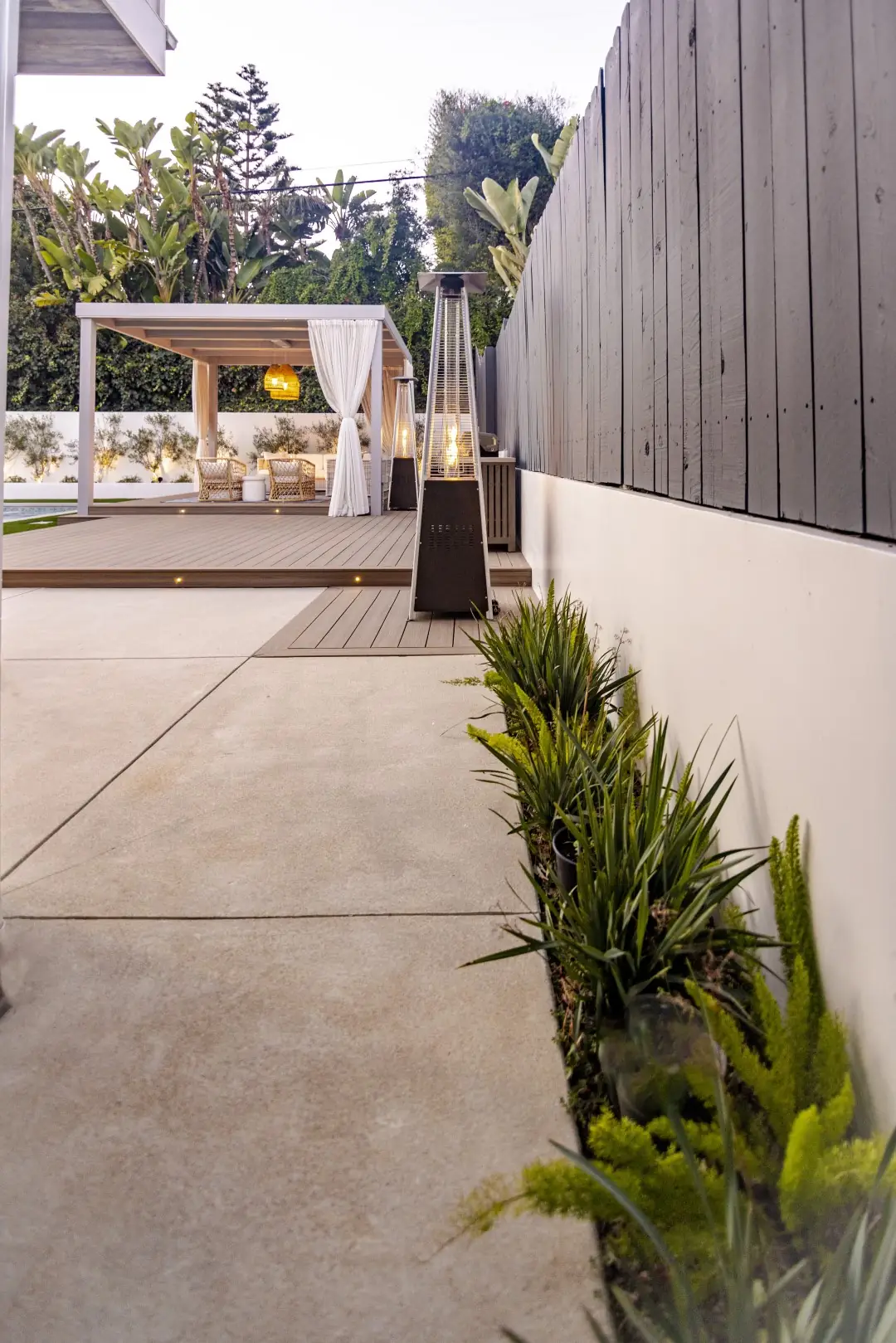 Concrete side yard path with drought-tolerant plantings along a dark wood fence, leading to a curtained cabana in Los Angeles