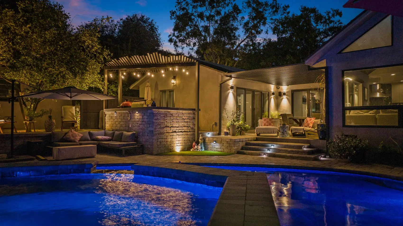 Paver patio with lounge seating and lit steps beyond the pool at night in Pasadena