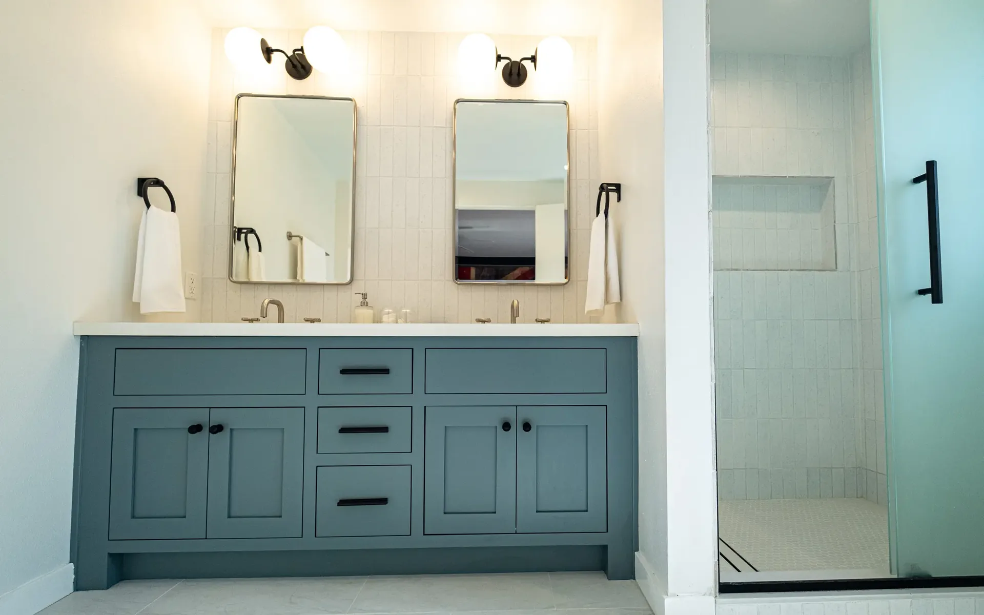 Remodeled Los Angeles bathroom with a teal double vanity, black hardware, twin mirrors with sconces and a frameless glass shower with a tiled niche.