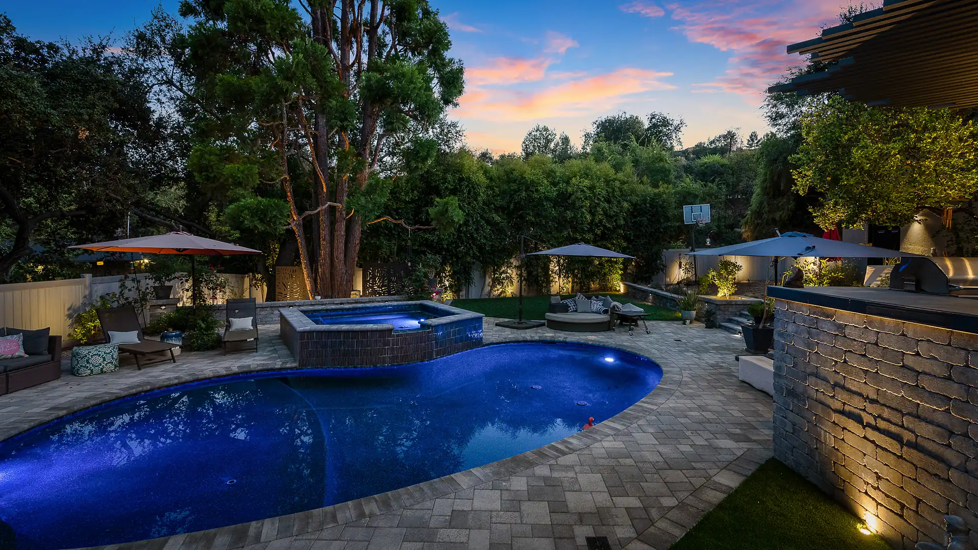 Luxury kidney-shaped pool glowing sapphire blue at dusk beside a raised stone-tiled spa, with paver decking, a stacked-stone wall with uplighting, cushioned lounge seating under patio umbrellas, and towering trees against a pink and blue sunset sky.