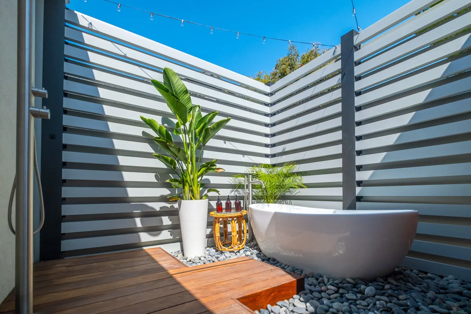 Freestanding soaking tub on a pebble bed with a wood platform and outdoor shower inside a slatted enclosure in Los Feliz