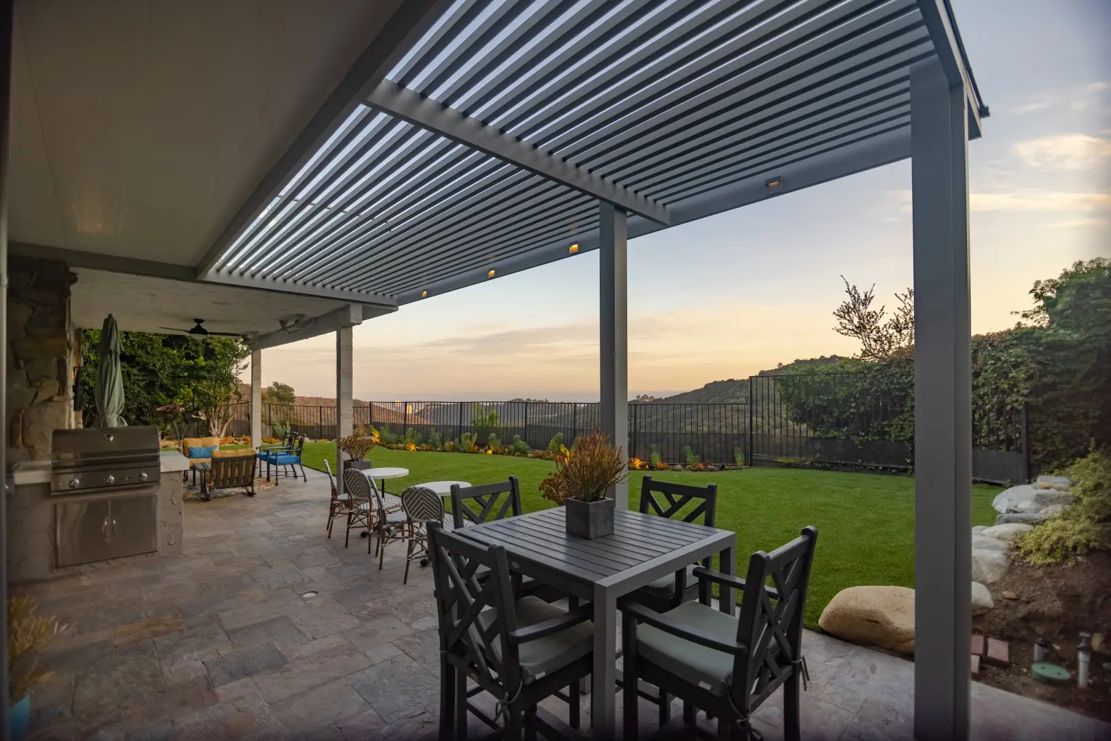 Louvered aluminum pergola over an outdoor kitchen and dining terrace at dusk with hillside views in Beverly Glen, Los Angeles
